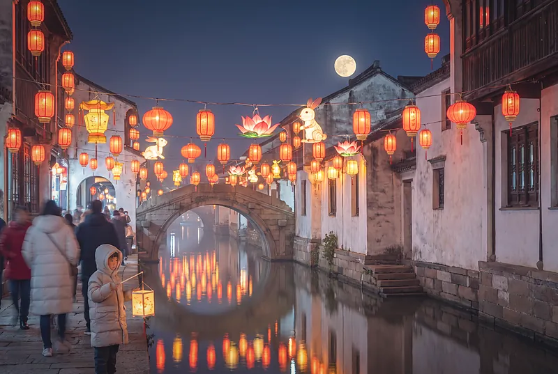 古镇元宵灯会夜景,河边挂满红色与金色灯笼,人群在灯下游赏
