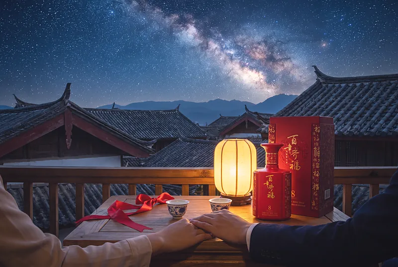 夜晚阳台上,一对恋人的手放在桌上,两只小酒杯和一条红色丝带,头顶是璀璨银河与中式屋檐