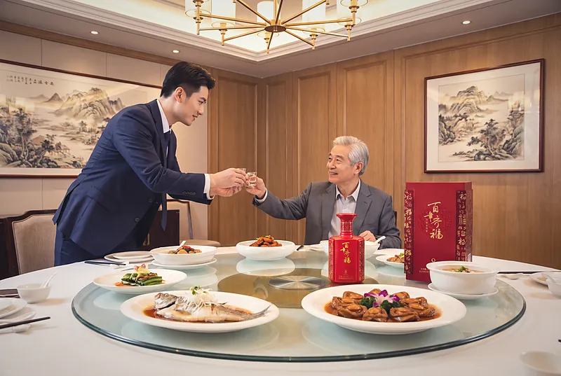 正式的中式商务宴席上,一位宾客在圆桌旁双手举杯向长者敬酒,桌上摆着百年齐福·8